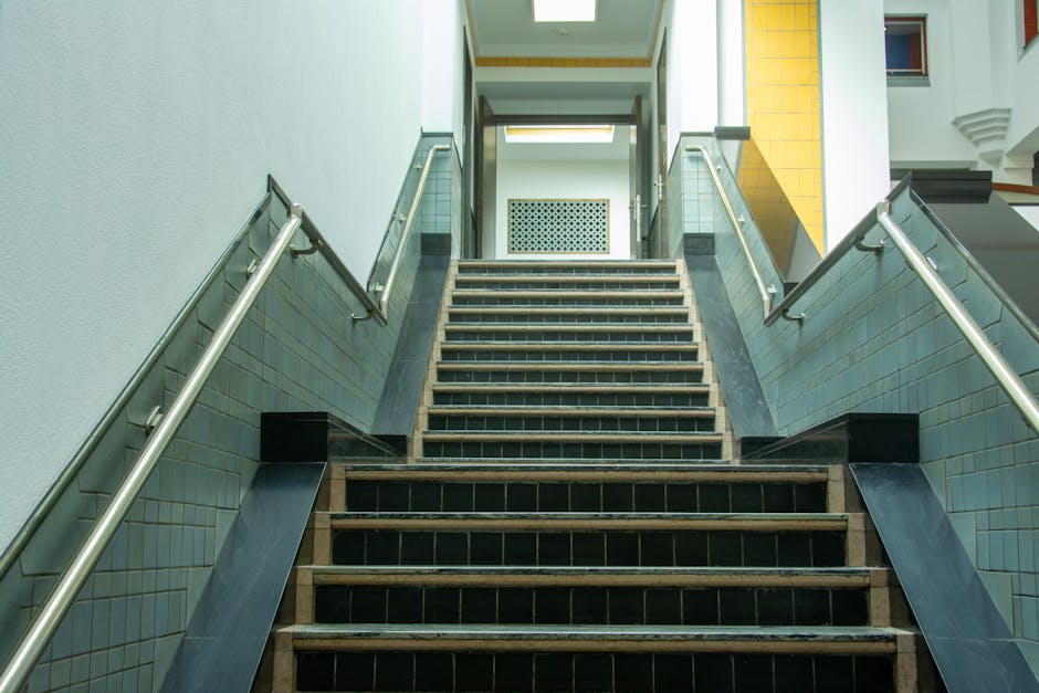A section of indoor staircase with black steps and a wooden handrail on the right side, attached to a white tiled wall. The staircase appears narrow, with some steps showing signs of wear and dirt. A