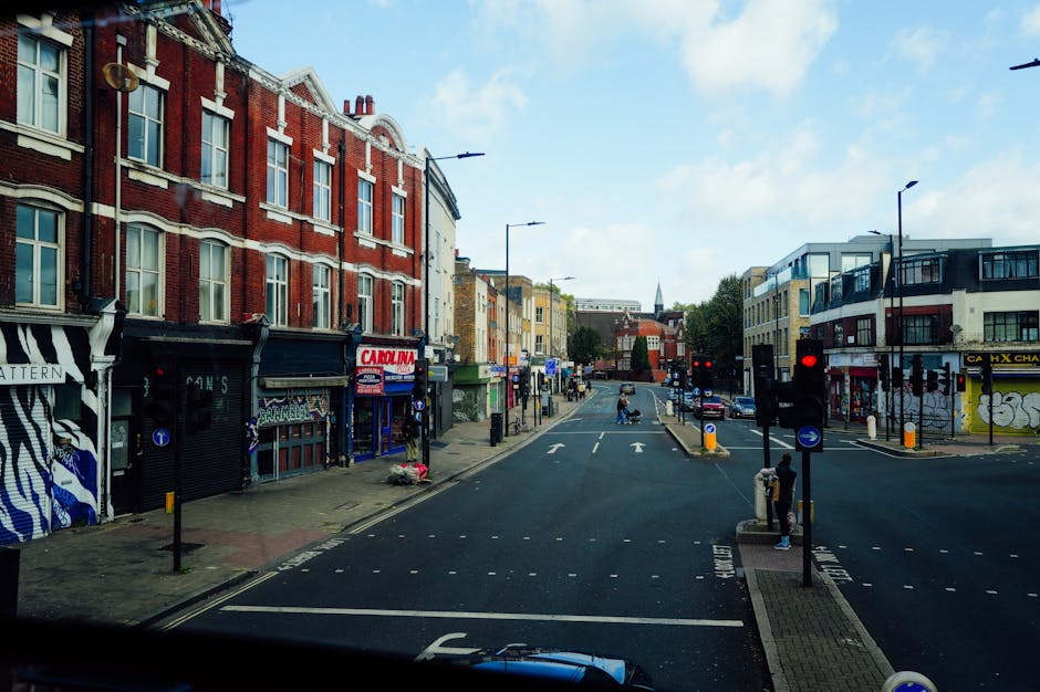 View of a city street in Elephant and Castle during daytime, showing a row of red brick and modern commercial buildings lining both sides of the road. The street features traffic lights, pedestrian crossings, and a few pedestrians waiting to cross or walking along the pavements. Several boxes and wrapped furniture pieces are visible on the pavement, indicating preparations for a home relocation or furniture transport. A van is partially visible at the bottom of the image, with some items being loaded or unloaded near the entrance of a building, supported by the professional removals services of manwithvanelephantandcastle.co.uk. The scene is illuminated by natural daylight with a partly cloudy sky overhead, and the environment reflects typical urban moving logistics including furniture packaging, loading, and street planning for efficient transport.