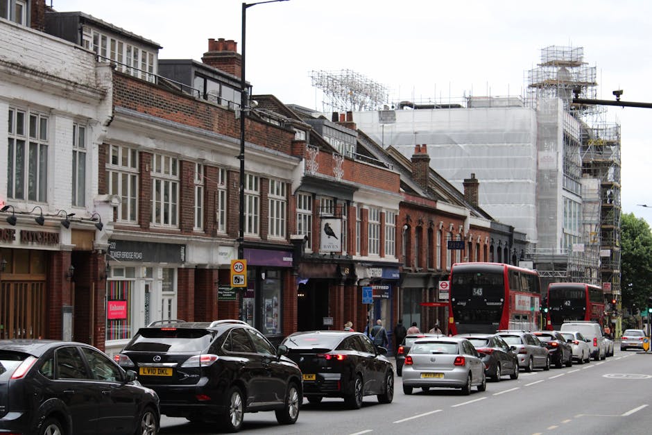 A street scene on Walworth Road near Elephant and Castle featuring a row of small, brick-fronted commercial buildings with large front windows and signage, some of which display retail businesses. In front of the buildings, there is a single lane of parked cars, including a black SUV with an open trunk, with several other vehicles parked along the curb. On the roadway, multiple vehicles are in motion, including two red double-decker buses and several smaller vehicles. The sidewalk has pedestrians walking, and some are near the entrances of the shops. Behind the buildings, there is scaffolding and construction work visible on a taller structure wrapped in white protective sheeting, indicating ongoing building renovation or development. The scene is set during daytime with natural lighting, and the environment overall reflects urban city life, typical for a busy shopping and transport corridor near Elephant and Castle, with [COMPANY_NAME] providing professional house and furniture removals in the area.
