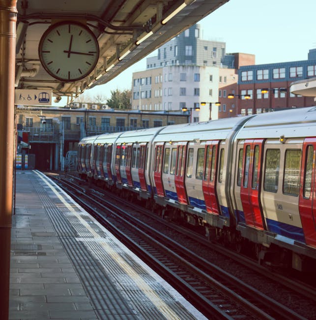 A London Underground train with a red, blue, and silver exterior is stationary on the tracks at an outdoor station near Elephant and Castle. The platform is partially covered by a roof, which features a large round clock showing the time as approximately 3:16. On the platform, there are tactile paving strips near the edge for accessibility and a few signs indicating station facilities. In the background, modern multi-storey buildings with a mix of glass and brick facades are visible under a clear sky. The station environment is clean and well-lit, with the train ready for passenger boarding or alighting, and the area appears to be in an urban setting experiencing daytime activity. This scene illustrates elements typical of urban transport infrastructure and is relevant to the context of house removals and moving logistics, such as transporting furniture or packing materials to a new home near Elephant and Castle.