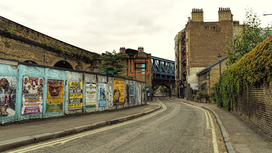 A narrow street near Elephant and Castle with a curved asphalt road featuring double yellow lines along the edges. On the left, a weathered metal fence is covered with colorful posters and advertisements, including event promotions and concert flyers. Behind the fence, part of a brick building with arched windows is visible, as well as a green, leafy plant climbing the fence. To the right, a tall brick wall with a dense hedge running alongside it lines the sidewalk. In the background, a blue metal railway bridge spanning the street is visible, connecting the buildings on either side. The scene is illuminated by natural daylight under an overcast sky, with no visible pedestrians or vehicles. This urban setting captures the typical environment where house removals and furniture transport may take place when preparing for home relocation near Elephant and Castle, supported by professional removals services such as those offered by manwithvanelephantandcastle.co.uk.
