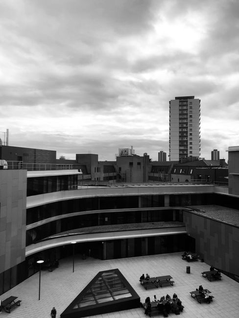 Black and white photograph of a modern urban outdoor space overlooking a cityscape with multiple high-rise buildings in the background. The foreground features a large courtyard or terrace area with several wooden benches, some occupied by small groups of people. The open space is paved and has a distinct geometric glass-roof pavilion at its center. Surrounding the courtyard are curved, multi-level structures with smooth, horizontal lines and large windows. The sky above is cloudy, contributing to the overall neutral lighting. This setting appears to be part of a contemporary residential or commercial complex, possibly associated with student housing or office buildings, and could be relevant in the context of house moves, furniture transport, or loading processes for relocation services, as described on manwithvanelephantandcastle.co.uk.